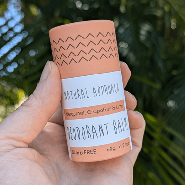 A hand holding a deodorant tube with natural approach branding, bicarb free, and 60g weight indication, against a backdrop of palm trees.